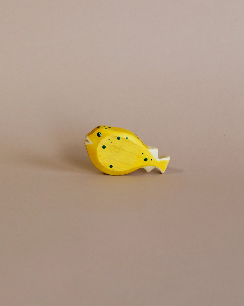 A small, yellow, wooden fish figurine with black spots and a white tail rests on a beige surface and background—part of the Handmade Holzwald Underwater World Set.