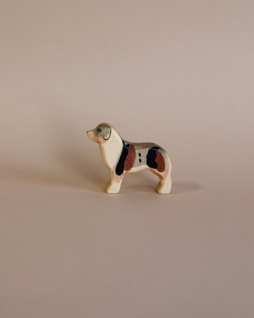 A Handmade Holzwald Australian Shepherd, painted with patches of black, white, and brown, standing against a plain light beige background. The dog appears in a stationary, profile pose.