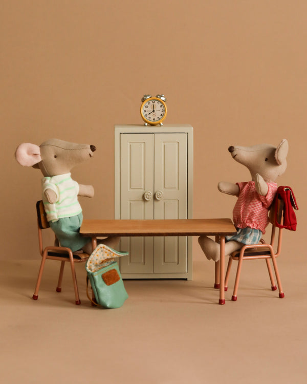 Two toy mice, dressed casually, sit on a wooden table with backpacks on the floor as part of the Maileg After School Set. A small cabinet with a clock is in the background, enhancing the charm of this quaint scene against a beige backdrop.