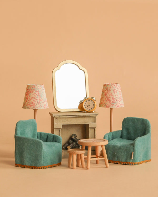 The Maileg Living Room Set - Mint features two teal armchairs, a fireplace with a mirror and alarm clock, two tables, and pink patterned lamps—ideal for displaying your Maileg mouse accessories against a beige background.