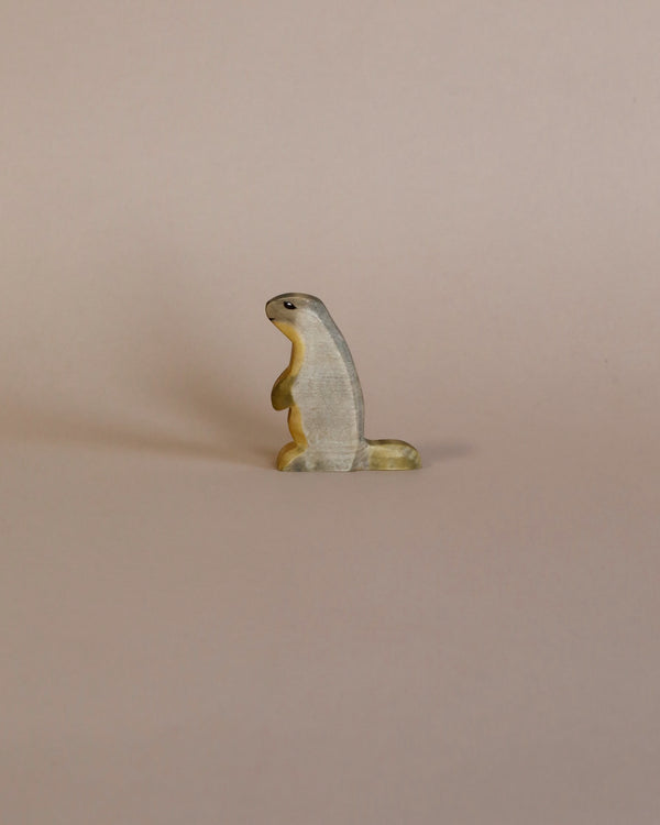 A small, solitary high-quality Handmade Holzwald Marmot figurine standing upright against a plain, light beige background. The figurine is textured and painted in natural colors.