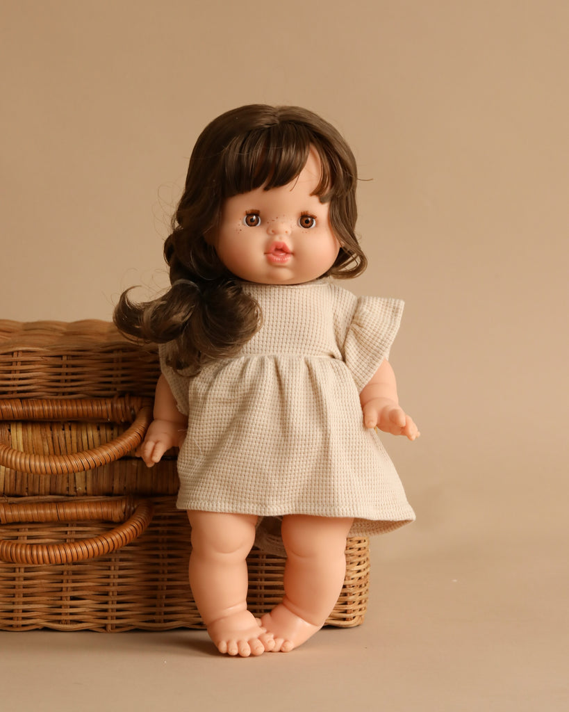 A Minikane Baby Doll (13") - Valentine with long brown hair stands next to a wicker basket against a beige background. The doll is wearing a simple beige dress, has a calm expression on its face, and features an anatomically correct design.