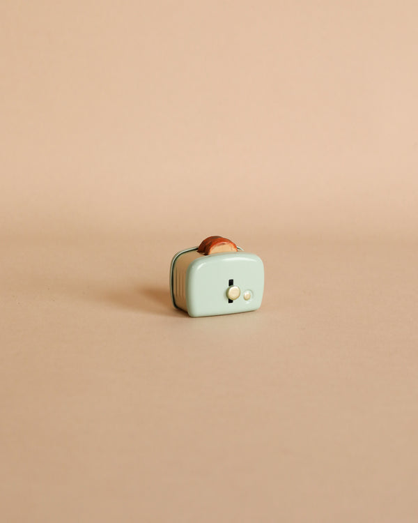 A small, quirky Maileg | Miniature Toaster - Mint kitchen timer with a piece of toasted bread sticking out the top, set against a plain light peach background.