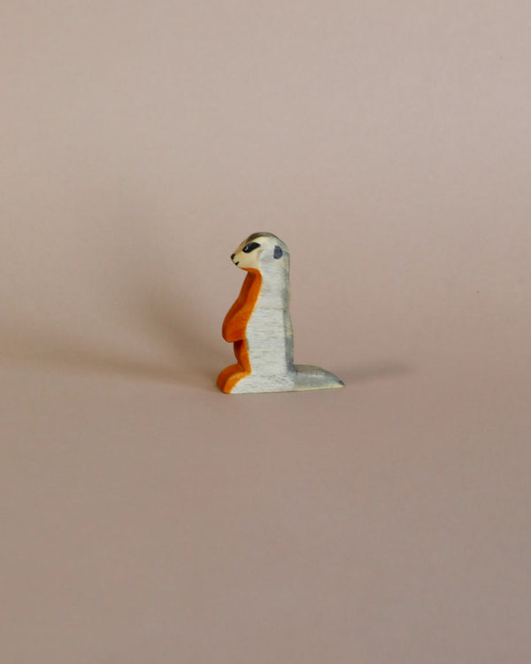 A small, whimsical sculpture of a Handmade Holzwald Meerkat with a white body and an orange beak, crafted with high-quality craftsmanship, set against a plain light pink background.