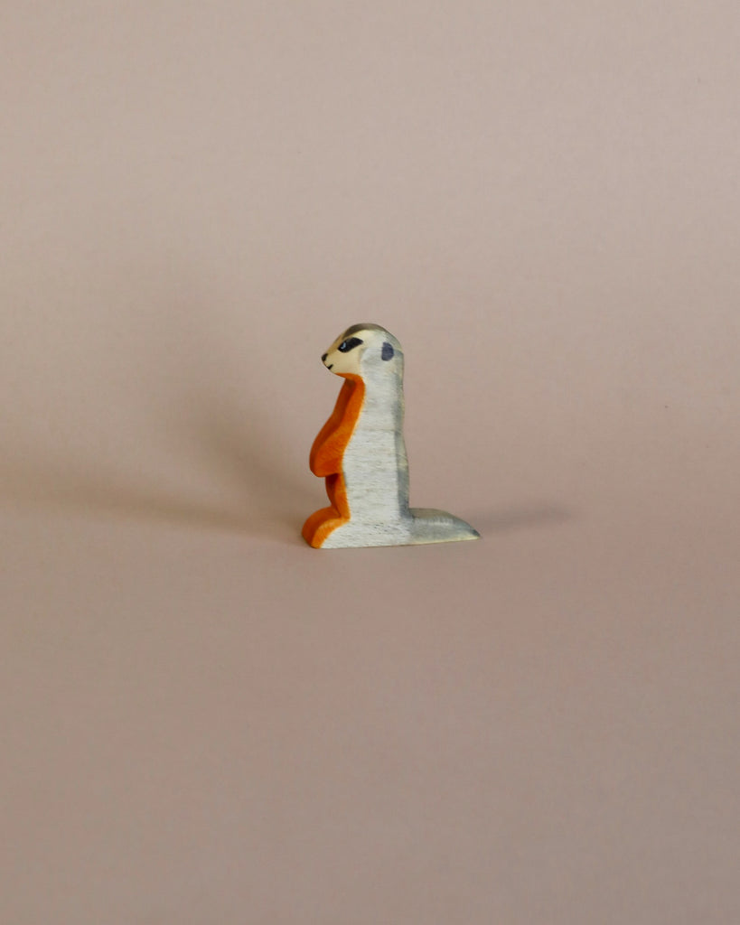A small, whimsical sculpture of a Handmade Holzwald Meerkat with a white body and an orange beak, crafted with high-quality craftsmanship, set against a plain light pink background.