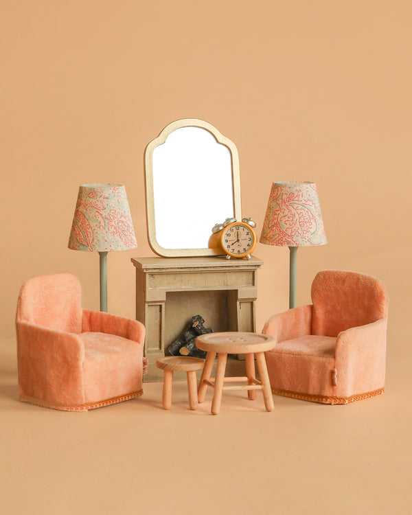 The Maileg Living Room Set includes two peach armchairs, a wooden table, two matching lamps, and a Maileg Fireplace with a mirror and clock, perfect for creating a charming miniature living room scene in your Maileg Dollhouse.