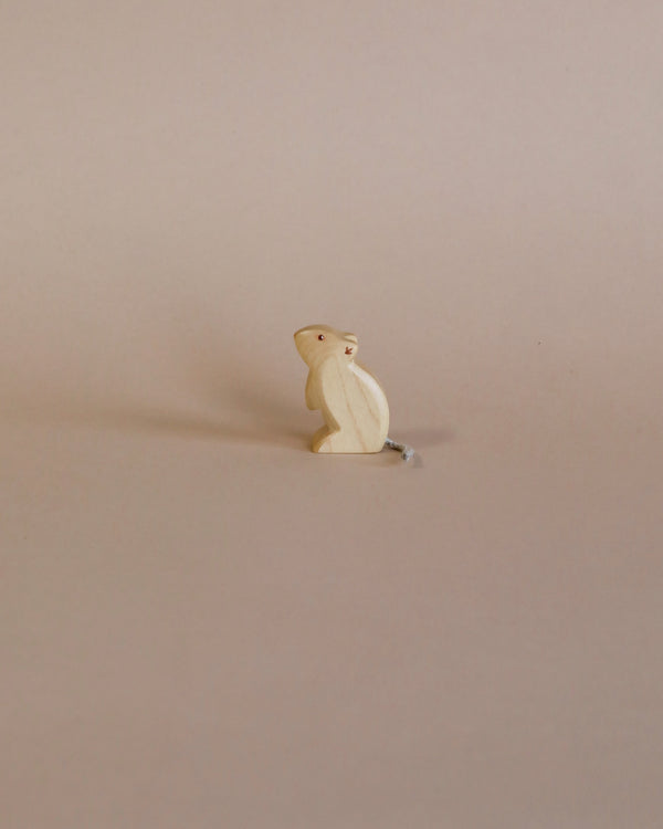 A small, simple wooden mouse figurine, crafted from sustainable toys materials, standing upright on a plain beige background, casting a soft shadow to its right.