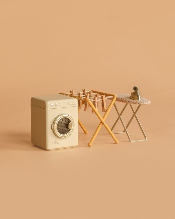The Maileg Laundry Room Set, featuring a miniature washing machine, drying rack with tiny clothes, and a small ironing board with iron, is displayed on a peach-colored background.