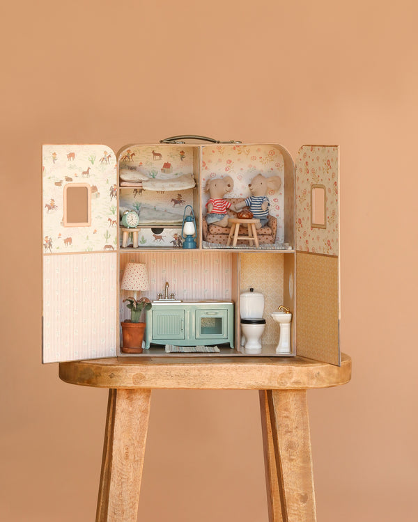 A Maileg Fully Furnished Suitcasa dollhouse with two rooms rests open on a stool. The left side has a kitchen and potted plant, the right is a bathroom, and Little Brother and Sister Mice sit in the cozy upper left bedroom.