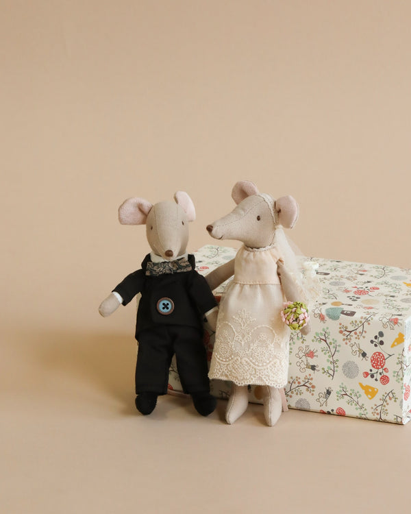 Two plush Maileg Wedding Mice Couple in Box dolls, dressed as a bride and groom, standing next to a floral gift box on a beige background. The groom mouse wears a black suit while the bride wears a lace dress.