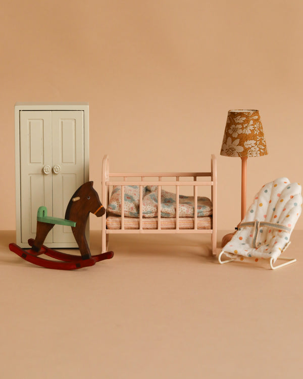 The Maileg Nursery Set on a beige background includes a white wardrobe, pink crib with bedding, brown rocking horse, floral vintage floor lamp, and polka-dotted baby bouncer—all sized for Maileg Mice.