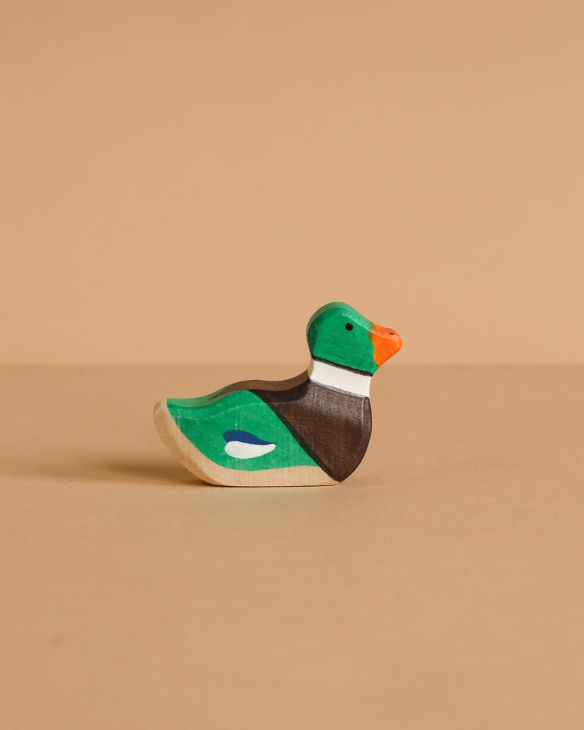 A Holztiger Handmade Wooden Woodland Animals Set features a small, handcrafted wooden duck figurine painted green, orange, brown, and white. It sits on a beige surface with a matching background—ideal for open-ended play.