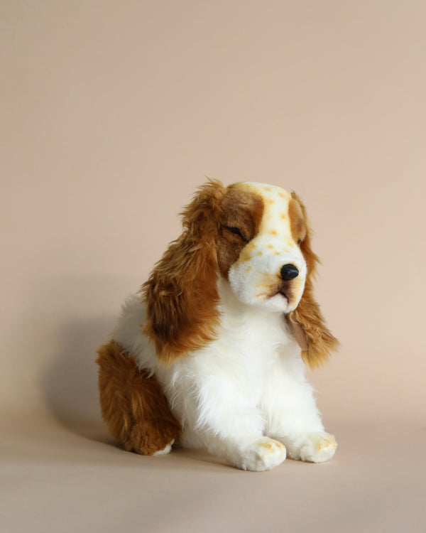 A Cocker Spaniel Pup Stuffed Animal, resembling a spaniel with brown and white fur and realistic features, seated against a plain beige background.