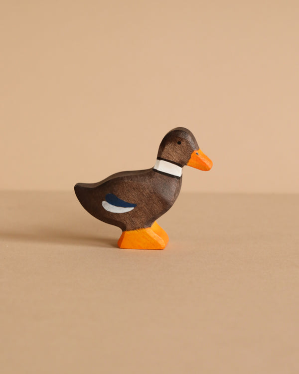The Holztiger Duck, Standing is a small, handcrafted wooden figurine painted brown with an orange beak and feet, a white collar, and a blue accent on its wing, shown on a beige surface with a beige background.