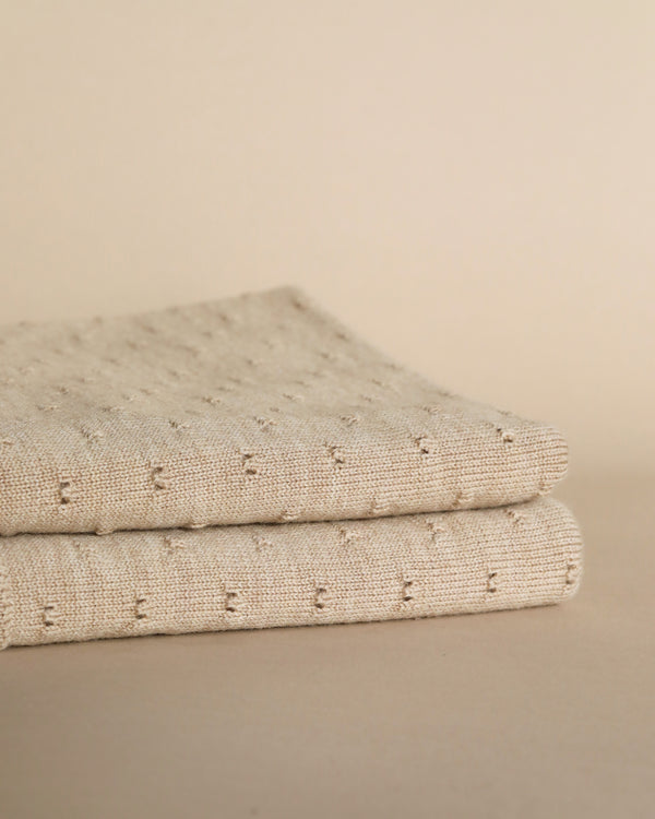 A folded Sand Merino Wool Bibi Blanket with delicate ajour embroidery on a soft beige background, showcasing a subtle texture and elegant pattern.