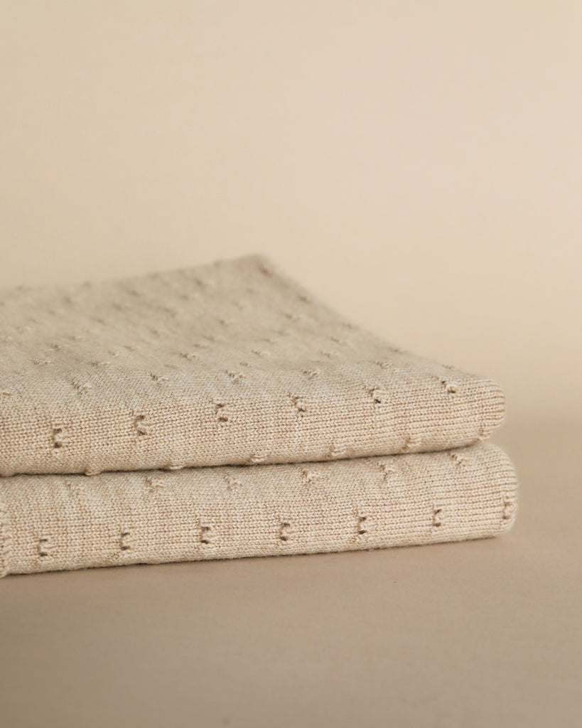 A folded Sand Merino Wool Bibi Blanket with delicate ajour embroidery on a soft beige background, showcasing a subtle texture and elegant pattern.