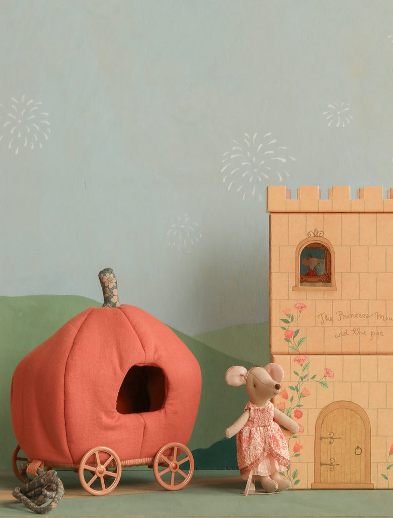 The Maileg Princess and The Pea, Big Sister Mouse in a pink dress stands beside a plush pumpkin carriage with wheels and a fairytale cardboard castle playset, set against a pastel backdrop with fireworks and green hills.