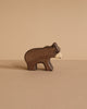 A small, dark brown handcrafted bear from the Holztiger Handmade Wooden Woodland Animals Set stands on a beige surface, featuring a white snout and classic design—ideal for open-ended play and reminiscent of iconic HOLZTIGER figures.