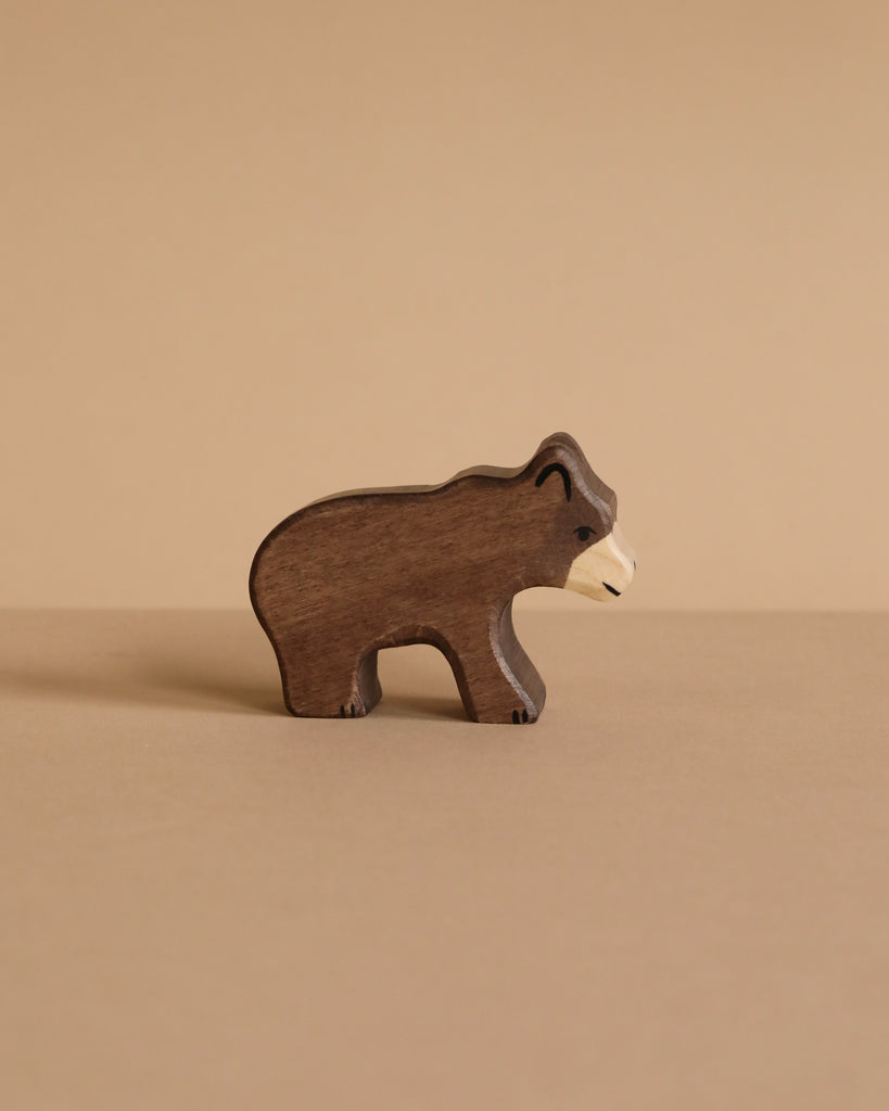 A small, dark brown handcrafted bear from the Holztiger Handmade Wooden Woodland Animals Set stands on a beige surface, featuring a white snout and classic design—ideal for open-ended play and reminiscent of iconic HOLZTIGER figures.