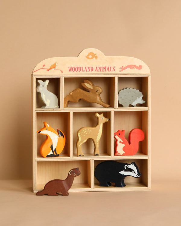 Sentence with Product Name: A wooden shelf labeled "Woodland Animals Set," containing painted wooden figures of various animals including a fox, deer, badger, and others against a beige background. Suitable for children age range 3 years