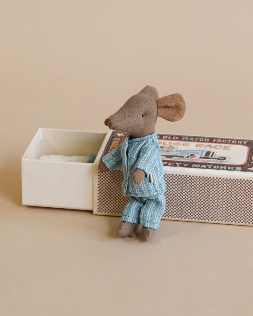 A small Maileg Big Brother Mouse in Box dressed in blue pajamas stands beside an open matchbox bed with pillows, against a tan background.
