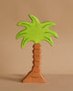 A handcrafted wooden palm tree from the Holztiger Handmade Wooden Safari Animals Set, featuring a light brown trunk and vivid green leaves, stands upright against a beige background—perfect for imaginative play or collection.