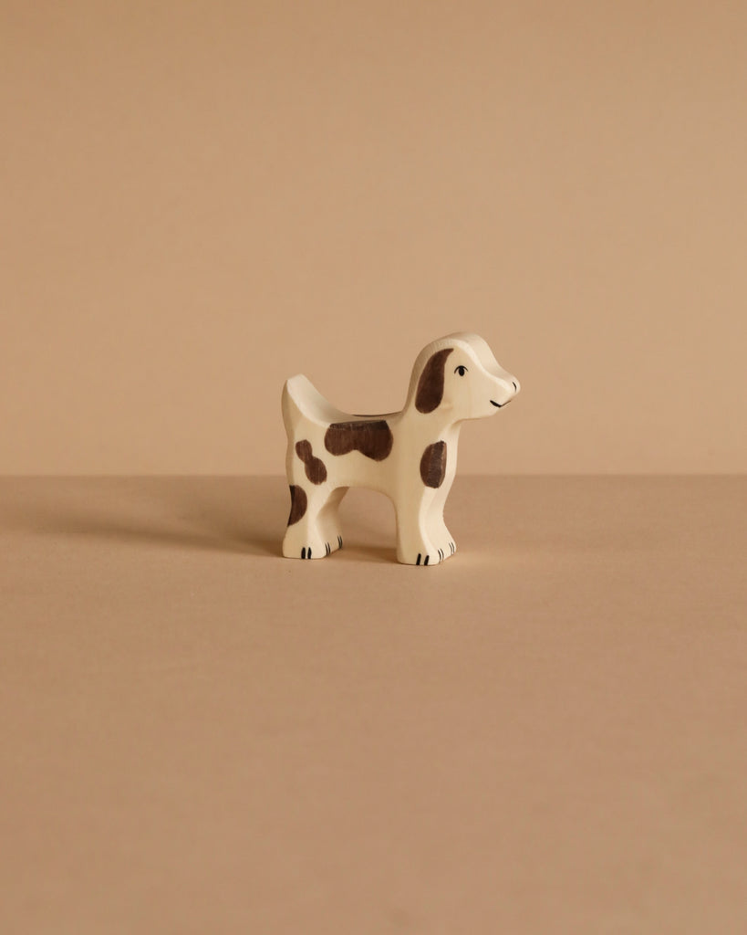 A Holztiger Small Farm Dog, a handcrafted white wooden figurine with brown spots, stands on a beige surface against a matching background, highlighting its high-quality artistry.