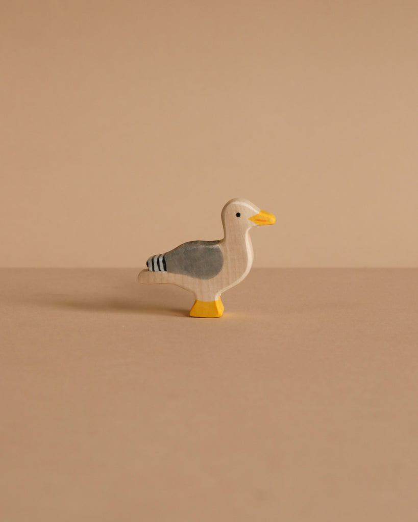 The Holztiger Seagull is a small, handcrafted wooden figurine featuring gray wings, a white head, yellow beak and feet. It stands on a plain beige surface with a matching background. Made in Europe by HOLZTIGER.