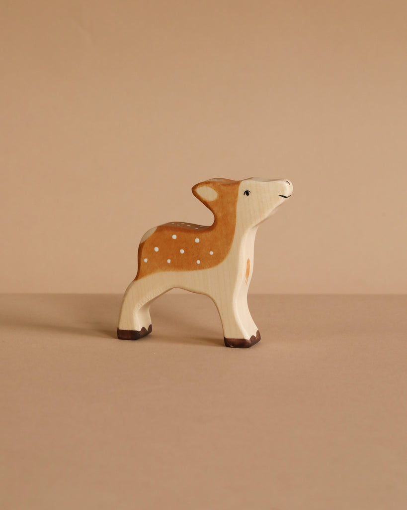 The Holztiger Fawn, a small handcrafted wooden deer with brown and white painted details, stands on a beige surface against a matching beige background.
