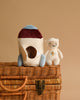 The Teddy In Outer Space Stuffed Animal - Rocket and a smiling teddy bear plush stand on top of a wicker basket. The rocket is primarily white with blue and maroon accents, while the teddy bear is off-white with a small embroidered emblem on its chest. The background is plain beige, setting the stage for endless space adventures.