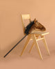 A wooden chair against a beige background holds the Pretend Play Ride-On Horse, featuring a plush horse head and black wooden stick, draped over the seat and resting on the floor—perfect for sparking imaginative play.