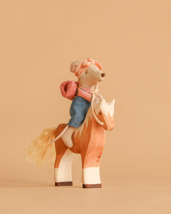 The Maileg Big Sister Horseback Adventure Set features a hiker mouse in denim pants, headscarf, and pink backpack riding a handmade wooden horse—an adorable scene perfect for fans of artisanal toys.
