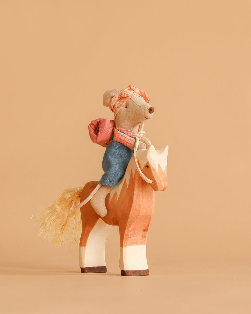 The Maileg Big Sister Horseback Adventure Set features a hiker mouse in denim pants, headscarf, and pink backpack riding a handmade wooden horse—an adorable scene perfect for fans of artisanal toys.