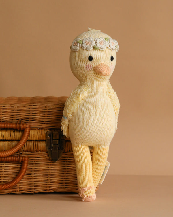 Cuddle + Kind Flora the Duckling - Ivory, a hand-knit duck with floral crown, leans on a wicker basket against a beige background—perfect for anyone seeking heirloom quality dolls.