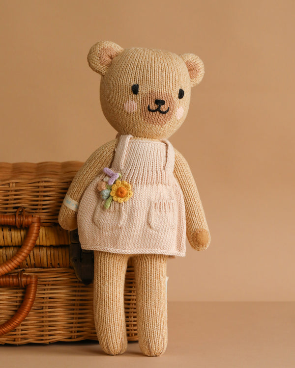 Cuddle + Kind Goldie The Honey Bear is a hand-knit teddy bear with a beige body and a cream apron dress adorned with a small yellow flower, pictured in front of a wicker basket on a plain tan background.