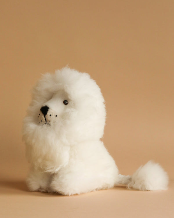 The Fluffy White Lion Stuffed Animal, sitting against a plain beige background, is a small, fluffy toy resembling a white lion. It features a mane and tail tuft made of soft alpaca wool, with black eyes and a black nose that give it an adorable, endearing expression.