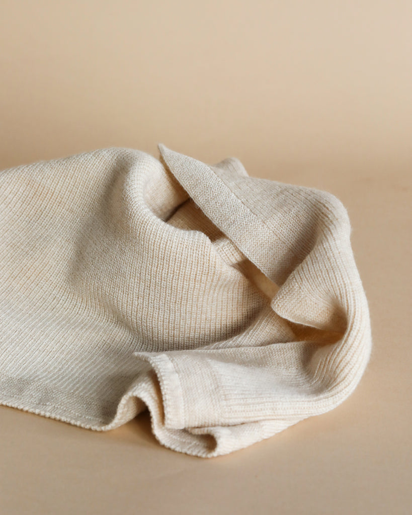 A crumpled Handmade Merino Wool Felix Blanket - Cream rests on a plain surface, illuminated by soft lighting, highlighting its texture and folds.