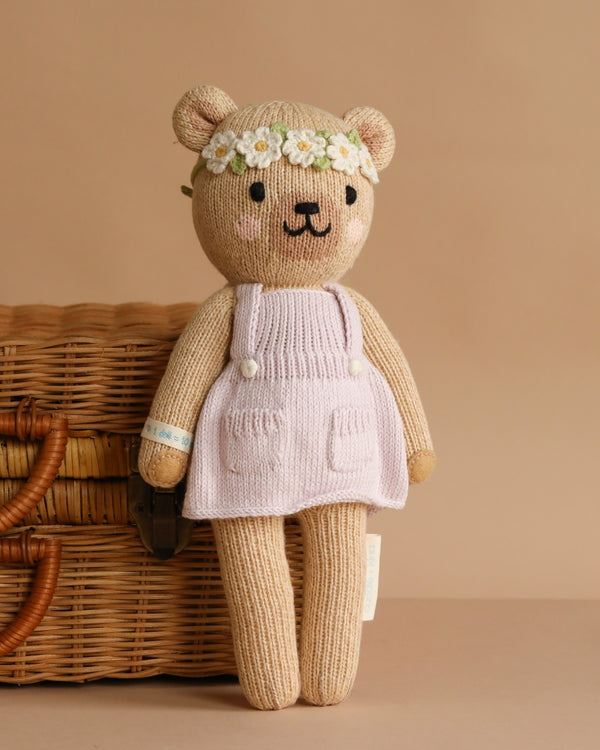 Cuddle + Kind Olivia The Honey Bear, a hand-knit teddy in a white dress and daisy flower crown, stands before a wicker basket against a beige background.