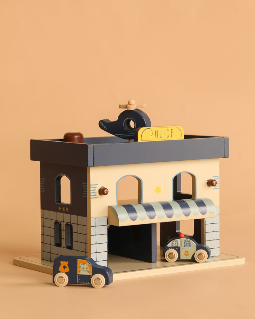 The Konges Sloejd Wooden Police Station features a blue and beige FSC-certified wood building with a POLICE sign, two toy cars, and a helicopter on the roof, all displayed against a plain tan background.