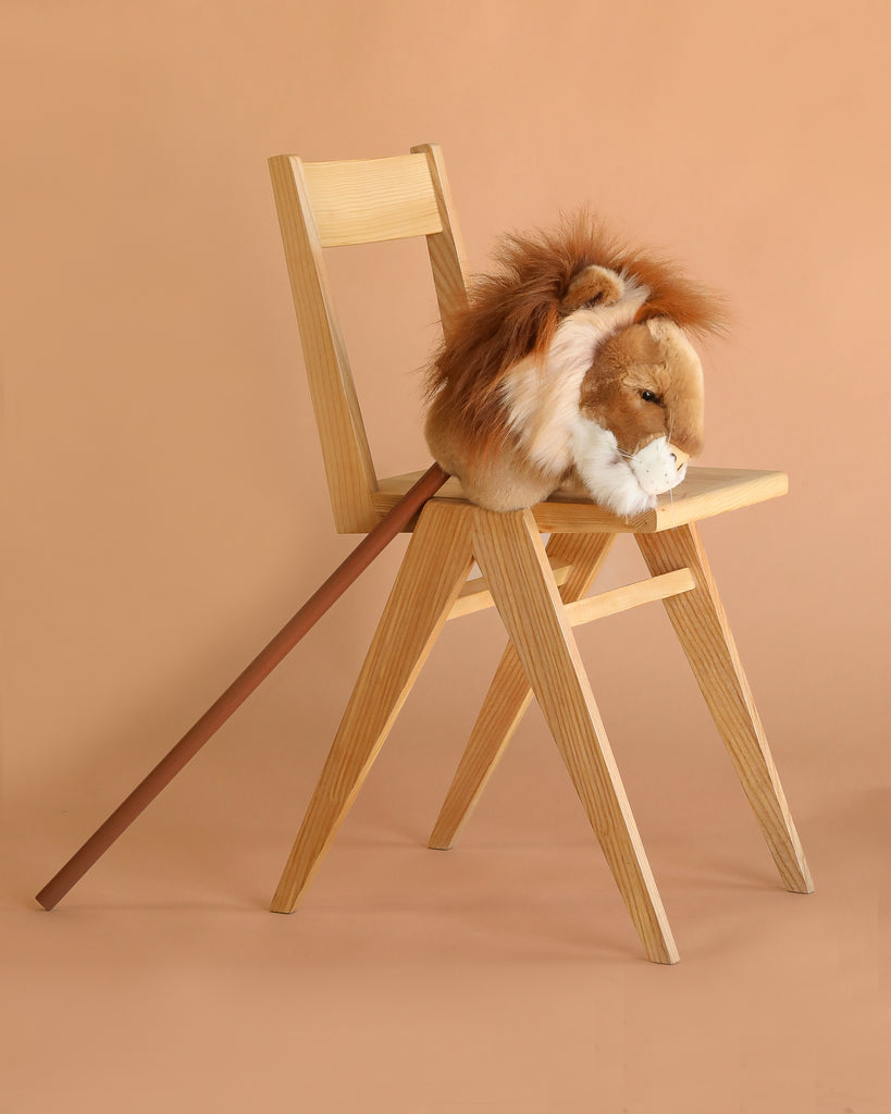 A wooden chair displays the Hobbyhorse - Lion, featuring a plush lion head and brown stick, ready for safari play against a simple beige background.
