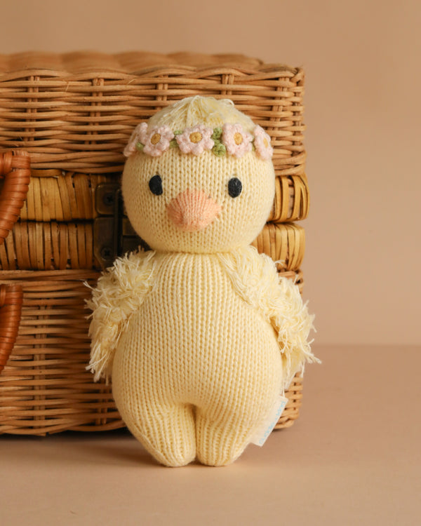 The Cuddle + Kind Baby Duckling, Blush Floral is a hand-knit yellow duck plush toy with a floral headband, posed in front of a wicker basket on a beige background.