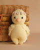 The Cuddle + Kind Baby Duckling, Blush Floral is a hand-knit yellow duck plush toy with a floral headband, posed in front of a wicker basket on a beige background.