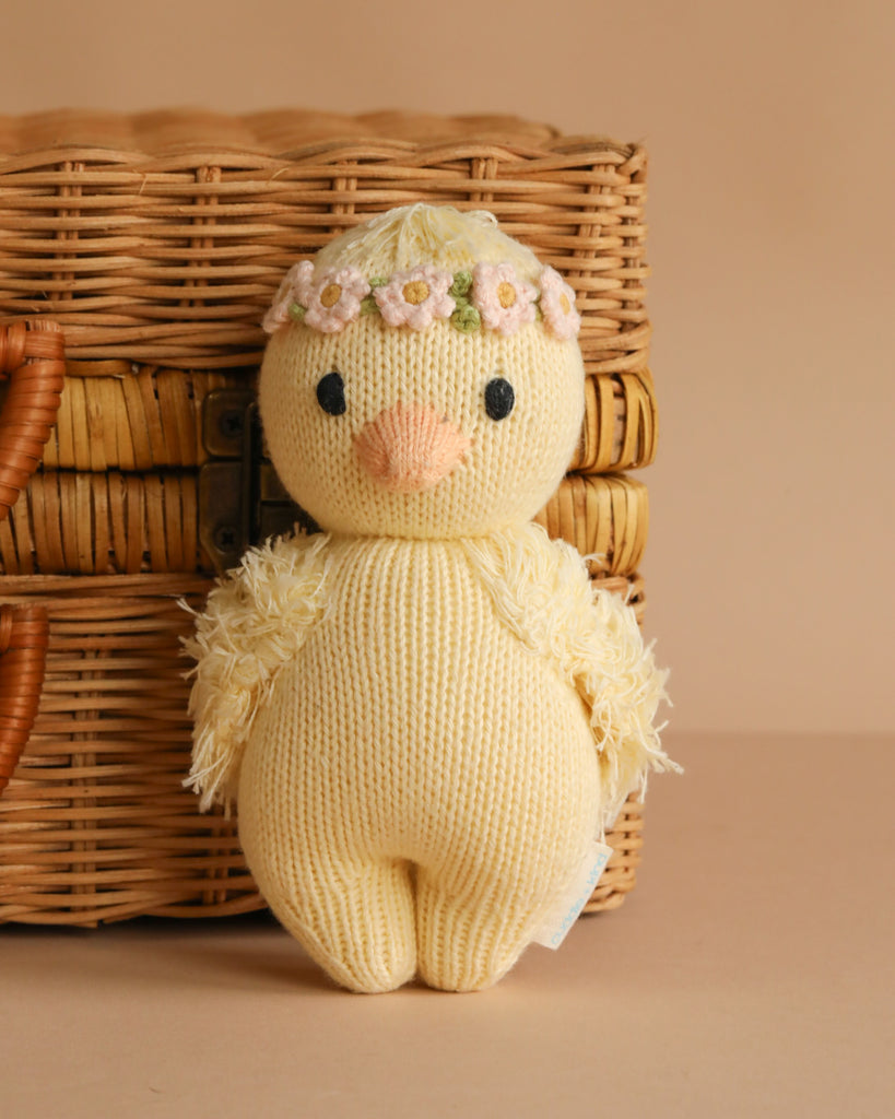 The Cuddle + Kind Baby Duckling, Blush Floral is a hand-knit yellow duck plush toy with a floral headband, posed in front of a wicker basket on a beige background.