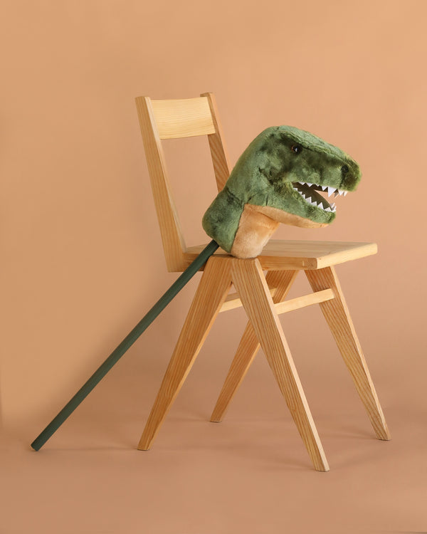 A wooden chair holds the Hobbyhorse - T-Rex Dino, a plush green dinosaur on a stick, perfect for imaginative play and standing out against a solid light brown background.