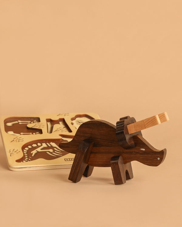 A Handmade Wooden Paleo Triceratops Dinosaur stands assembled in the foreground on a beige surface, while an animal-shaped wooden puzzle is visible in the background.