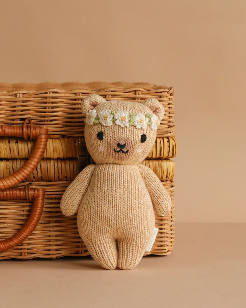 The Cuddle + Kind Baby Honey Bear, Ivory Floral—a hand-knit teddy bear crafted from soft Peruvian cotton yarn with a floral headband—sits in front of a woven wicker basket on a plain beige background.