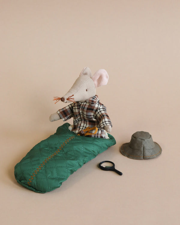 A Maileg Wildlife Guide Mouse dressed in a plaid shirt and vest, sitting on a green sleeping bag next to a gray hat and a black magnifying glass, on a beige background.
