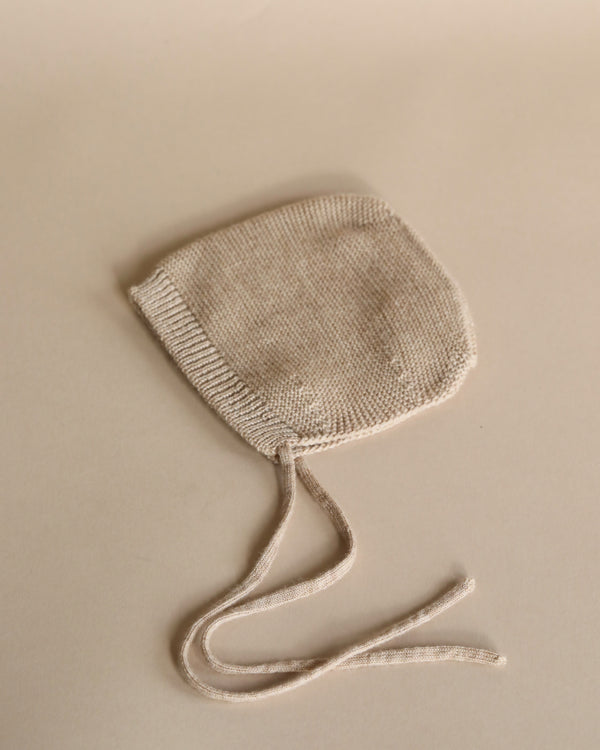 A small, knitted Sand merino wool baby bonnet with two long strings, laid flat on a beige surface.