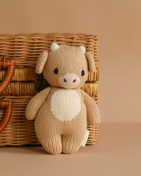 The Cuddle + Kind Baby Cow is a hand-knit beige plush made from heirloom-quality Peruvian cotton yarn, with white horns and a cream belly, pictured in front of a woven wicker basket on a light brown background.