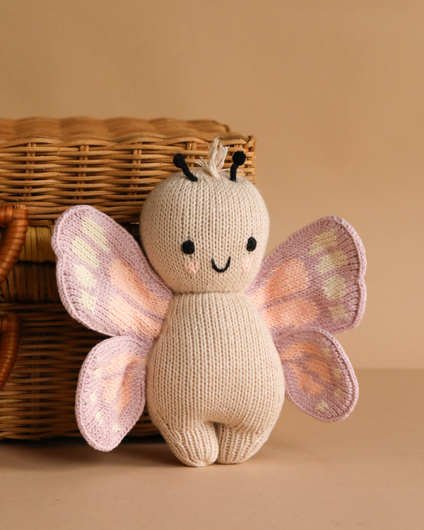 The Cuddle + Kind Baby Butterfly, a plush toy with light pink and purple wings and a smiling face, crafted from heirloom-quality Peruvian cotton yarn, sits in front of a wicker basket on a beige surface.
